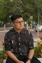 A man with glasses sits in a park wearing a patterned shirt and using earbuds.